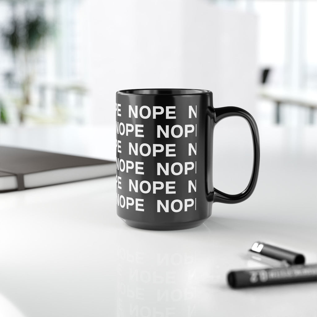A 15 oz black ceramic mug with a repeating white 'NOPE' text pattern, sitting on a white desk with a blurred notebook and pen in the background.
