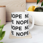 An 11 oz white ceramic mug printed with the word 'NOPE' repeated in bold black letters, sitting on a beige coaster on a white marble-like surface with a softly blurred kitchen background.