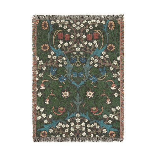 A rectangular woven blanket with fringed edges featuring John Henry Dearle’s  Blackthorn, depicting blackthorn blossoms, violets, fritillaries, daisies, twisting branches, and dense foliage together in a richly layered botanical pattern against a dark ground. Shown on a solid white background.