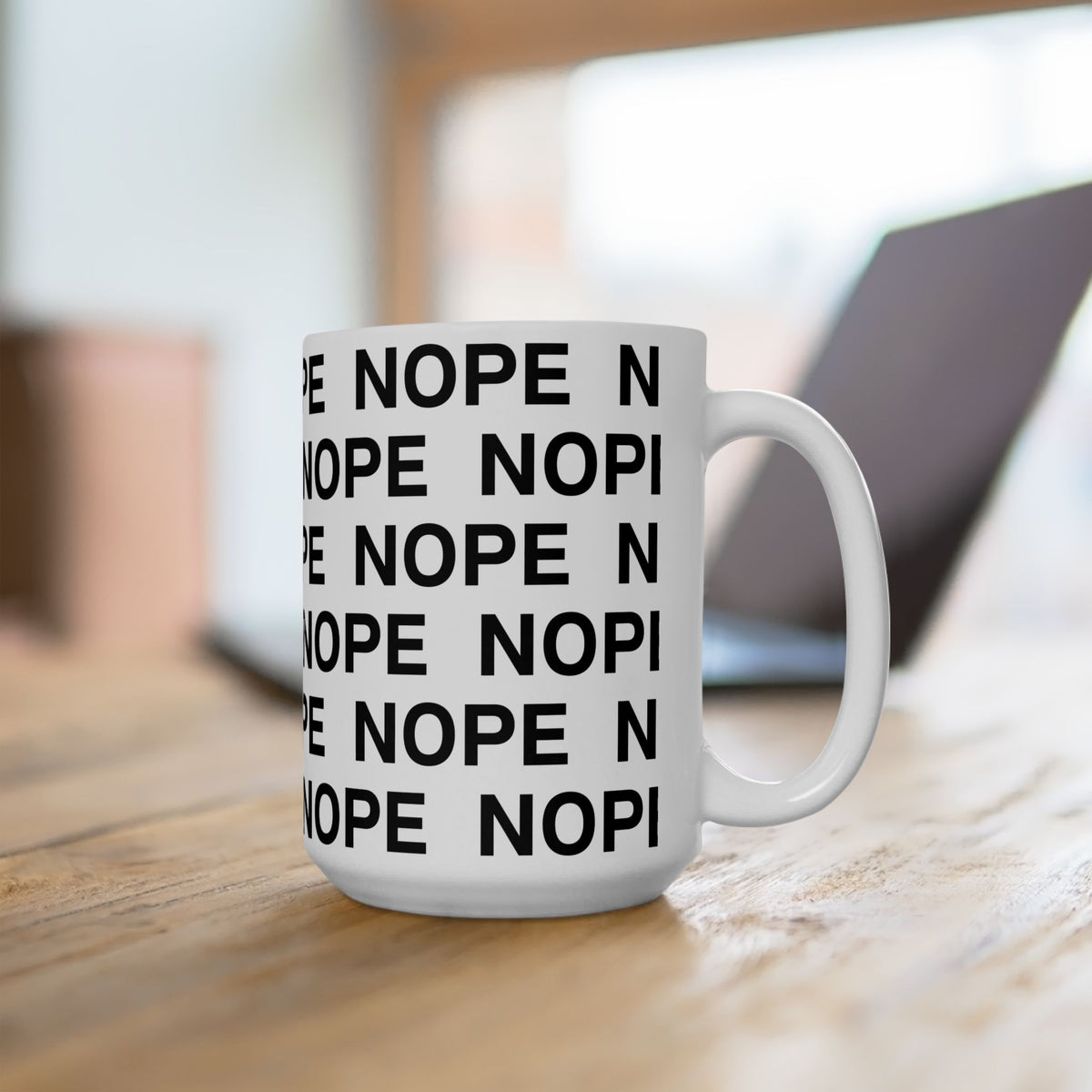 A 15 oz white ceramic coffee mug with the word 'NOPE' printed repeatedly in bold black capital letters, sitting on a wooden table with a blurred laptop in the background.
