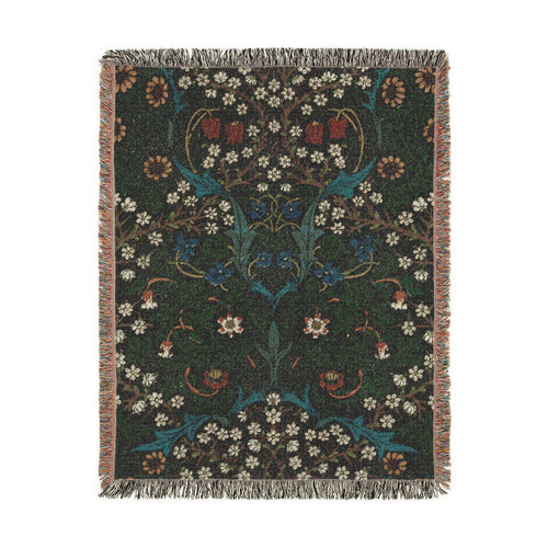 A rectangular woven blanket with fringed edges featuring John Henry Dearle’s  Blackthorn, depicting blackthorn blossoms, violets, fritillaries, daisies, twisting branches, and dense foliage together in a richly layered botanical pattern against a dark ground. Shown on a solid white background.