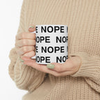 A person in a beige knit sweater holds an 11 oz white ceramic mug with the word 'NOPE' printed repeatedly in bold black capital letters.