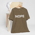 A heather olive crew neck t-shirt with 'NOPE' in bold white lettering across the chest on a wooden hanger and hung from the back of a wooden chair with a white hat also hanging from the chair. Shown against a plain white background.