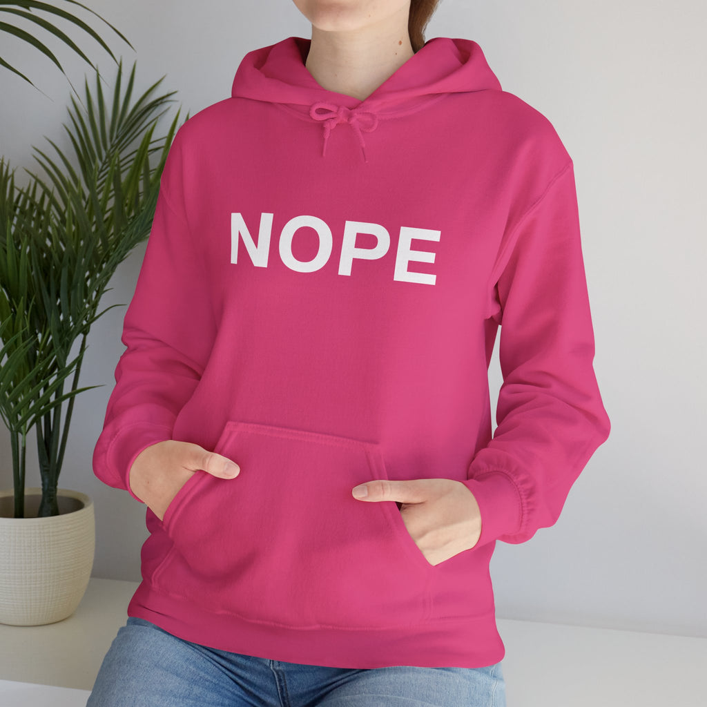 A person wearing a bright pink pullover hoodie with the word 'NOPE' printed in large white letters across the chest, paired with blue jeans, standing indoors near a potted plant.