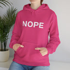 A person wearing a bright pink pullover hoodie with the word 'NOPE' printed in large white letters across the chest, paired with blue jeans, standing indoors near a potted plant.