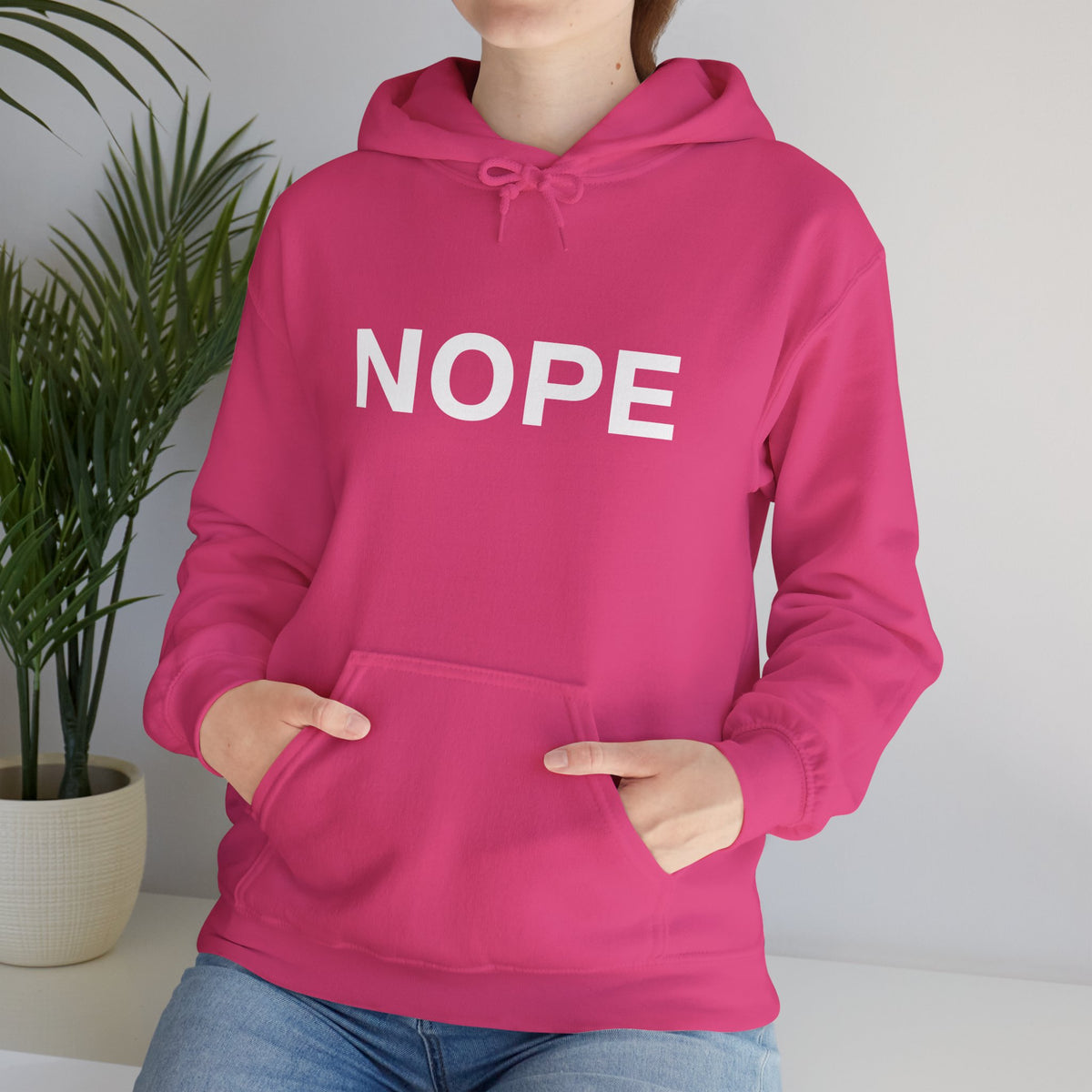 A person wearing a bright pink pullover hoodie with the word 'NOPE' printed in large white letters across the chest, paired with blue jeans, standing indoors near a potted plant.