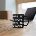 An 11oz black ceramic mug with the word 'NOPE' printed repeatedly in white block letters, sitting on a wooden table with a blurred laptop in the background.