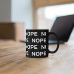 An 11oz black ceramic mug with the word 'NOPE' printed repeatedly in white block letters, sitting on a wooden table with a blurred laptop in the background.