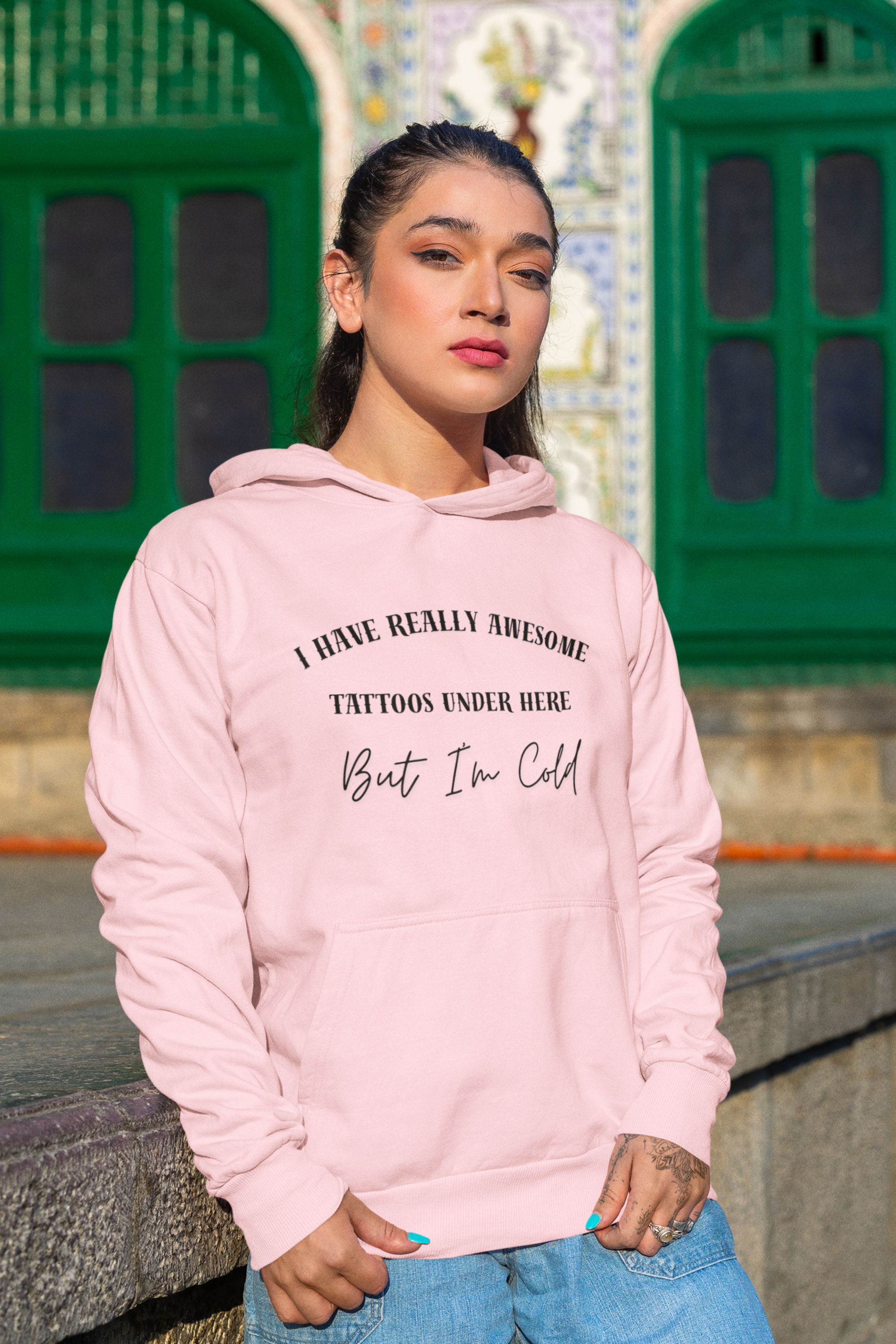 A woman wearing a light pink hoodie with black text that reads 'I HAVE REALLY AWESOME TATTOOS UNDER HERE But I'm Cold' in front of a stone wall and green arched doors.
