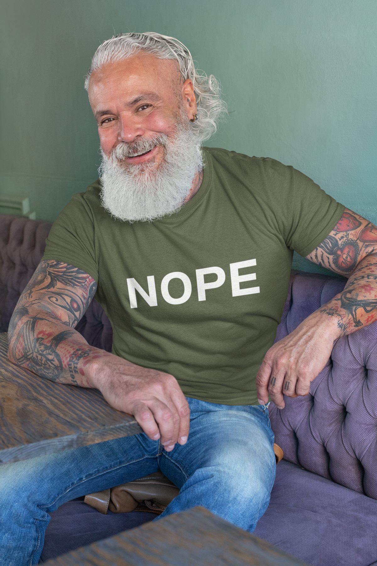 A smiling man with a white beard and tattoos sits on a purple tufted sofa wearing a military green t-shirt with the word 'NOPE' printed in bold white letters across the chest.