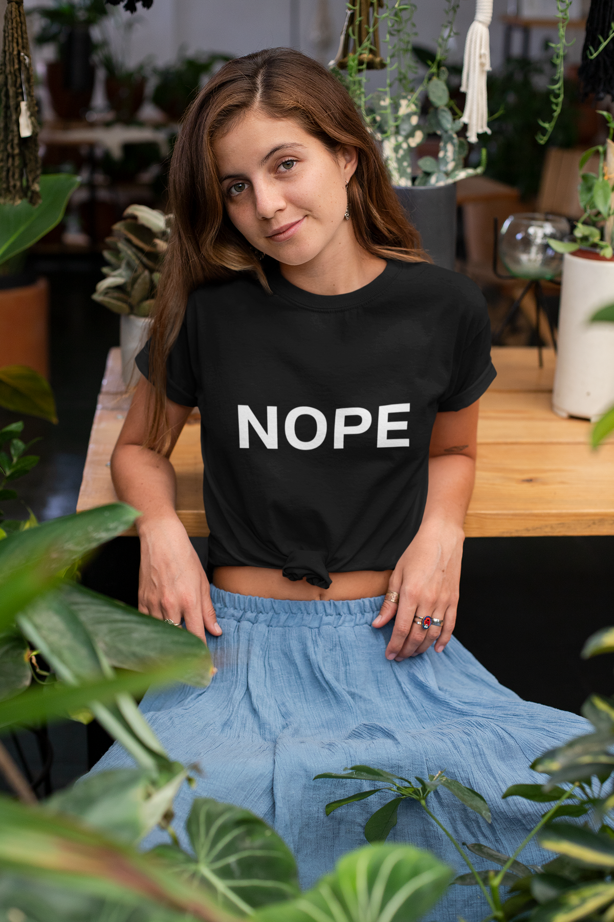 A woman wearing a black short-sleeve t-shirt knotted at the waist with the word 'NOPE' printed in bold white letters, paired with a light blue skirt, posed in a plant-filled indoor setting.