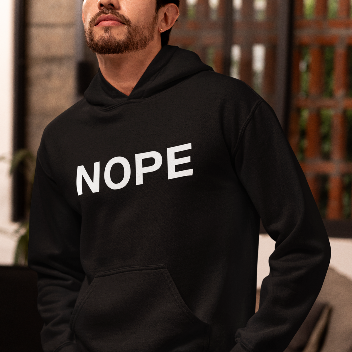 A man wearing a black pullover hoodie with the word 'NOPE' printed in bold white capital letters across the chest, photographed indoors with a softly blurred background.