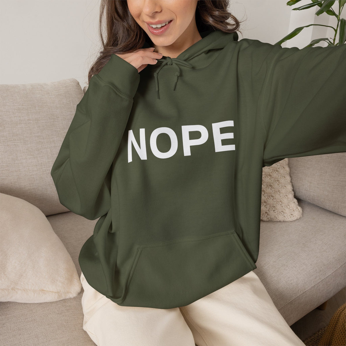 A woman wearing a military green pullover hoodie with the word 'NOPE' printed in large white letters across the chest, paired with white pants, sitting on a beige couch near a plant.