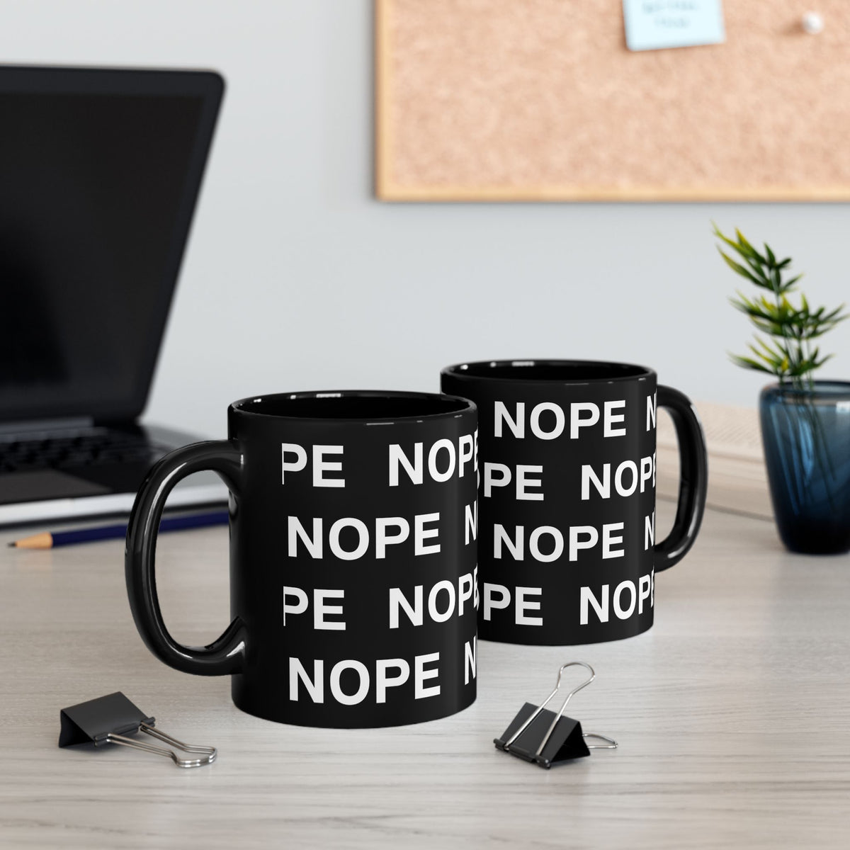 Two 11oz black ceramic mugs with white 'NOPE' text printed repeatedly, displayed on a light wood desk with office props including a laptop, binder clips, and a small plant.