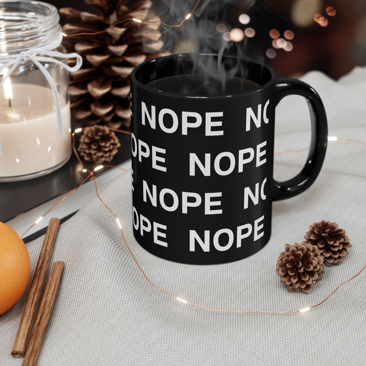 An 11oz black ceramic mug with a repeating white text graphic saying 'NOPE', filled with a steaming hot drink, styled on a light fabric surface with pinecones, a candle, cinnamon sticks, and fairy lights.